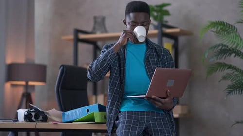 Selbstbewusster junger Afroamerikaner trinkt Morgenkaffee sitzend auf Tisch mit Laptop