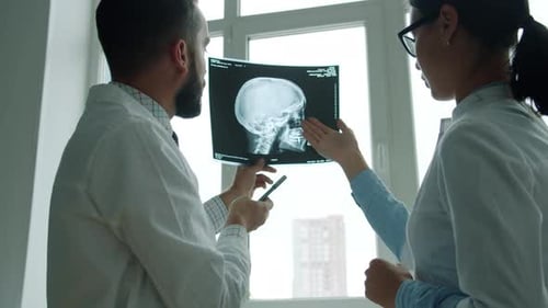 Girl and Guy in Doctor's Uniform Examining X-ray Images of Skull Talking in Clinic