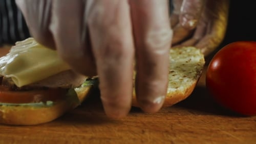 Gloved hands assembling sandwich with lettuce, tomato, cheese
