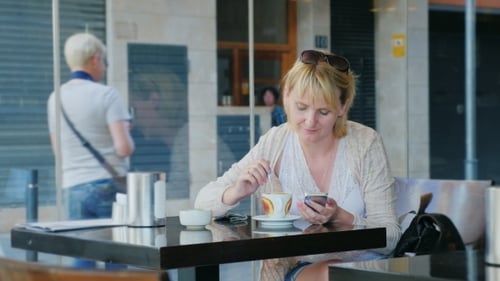 Young Woman Sitting By The Window In a Cafe. Is Typing On a Mobile Phone, Drinking Coffee. Outside