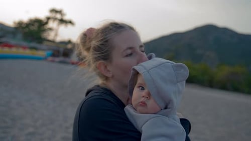 Mother Holds Cute Little Baby in Arms Standing on Beach