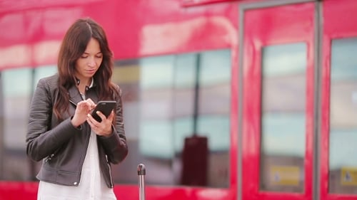 Junge Frau mit Gepäck schaut auf Handy an einem Bahnhof
