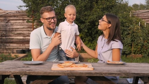 Young Happy Family Having Picnic In The Garden. Mom, Dad And Son Eating Pizza Outdoors