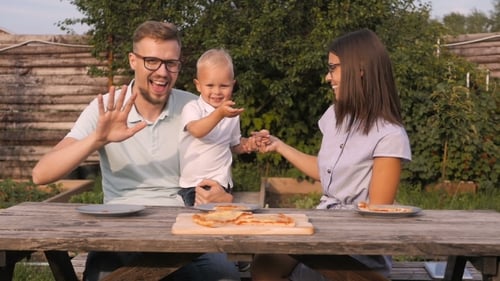 Young Happy Family Having Picnic In The Garden. Mom, Dad And Son Eating Pizza Outdoors