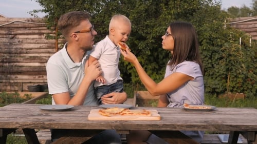 Young Happy Family Having Picnic In The Garden. Mom, Dad And Son Eating Pizza Outdoors