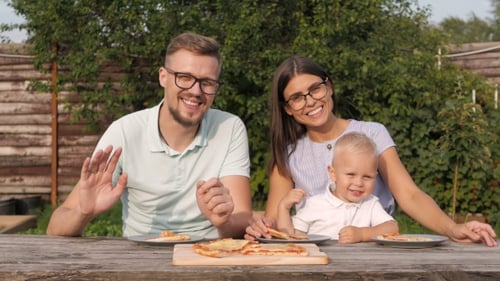 Young Happy Family Having Picnic In The Garden. Boy Waving At The Camera