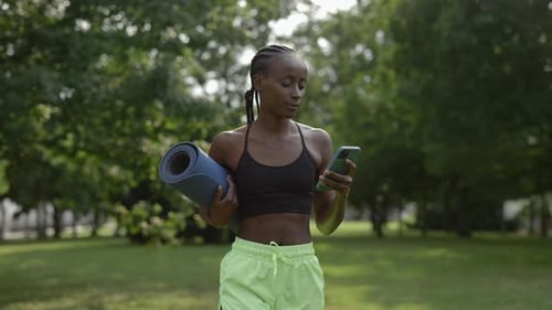 Woman in Sportswear Walking at Park with Mobile and Yoga Mat