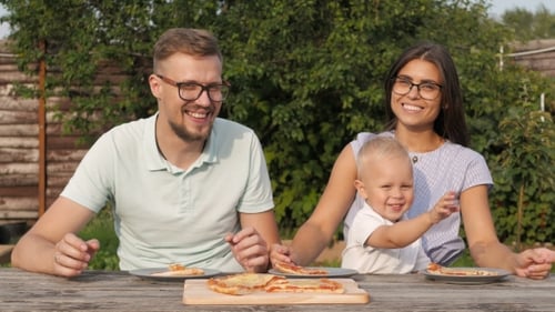 Young Happy Family Having Picnic In The Garden. Boy Waving At The Camera