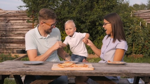 Young Happy Family Having Picnic In The Garden. Mom, Dad And Son Eating Pizza Outdoors