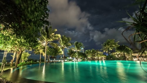 Wolken schweben über dem beleuchteten Nachtpool vor dem Hintergrund von Palmen und dem Meer.
-