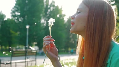 Young Woman Blowing Dandelion Seeds in Summer Park
