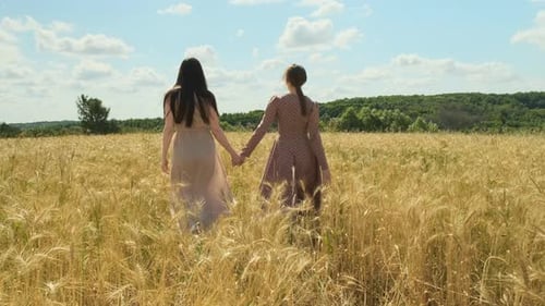 Two Enchanting Women Walking Holding Hands in Wheat Field in Beautiful Dresses and Looking at Each