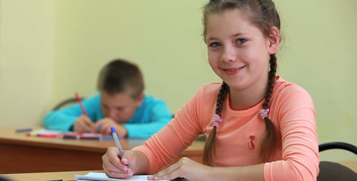 Young Girl Writing in Classroom with Boy in Background