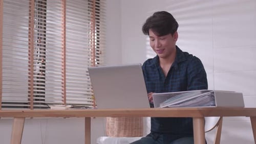 At home, a business asian man types and works online on a desk table with a laptop.
