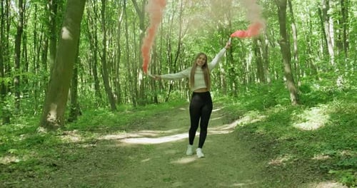 Beautiful Girl in a Green Forest Spins Around While Holding Two Red Smoke Bombs. Slow Motion