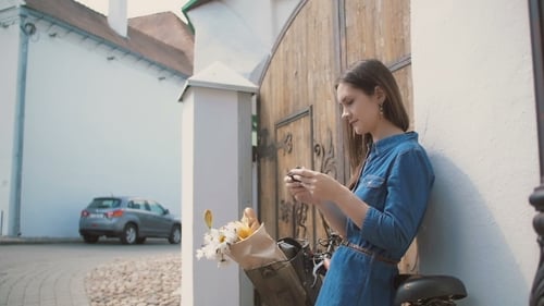 Brunette Girl Using Her Smartphone Standing Near Building With a Bike With Flowers In a Basket,
