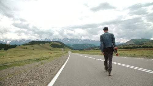 Man Carries a Longboard. The Boy Goes On The Asphalted Road Holding a Longboard