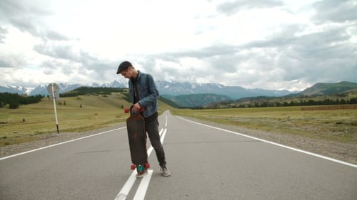 Man Carries a Longboard. The Boy Goes On The Asphalted Road Holding a Longboard