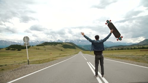 Man Carries a Longboard. The Boy Goes On The Asphalted Road Holding a Longboard