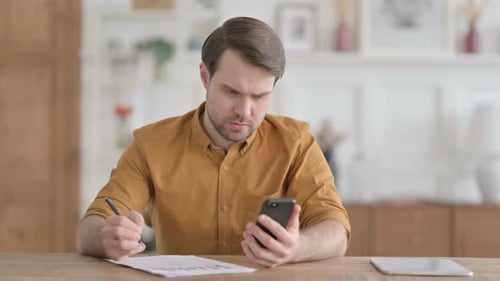 Junger Mann schaut aufs Telefon, während er im Büro auf Papier schreibt