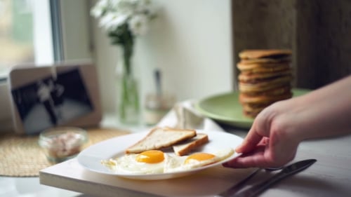Frau stellte Teller mit Spiegeleiern und Scheiben Toastbrot auf Küchentisch