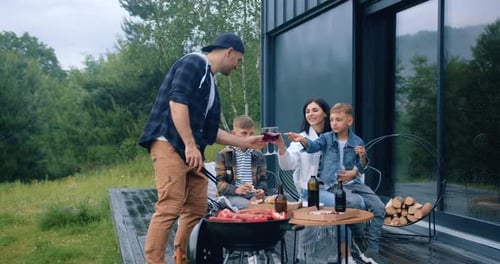 Father Sausages on Grill and Clinking Glasses with Wine with His Wife while Two Sons Outdoors