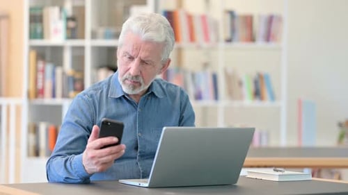 Senior Man Uses Smartphone at Laptop