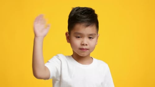 Little Asian Boy Waving Hand Looking At Camera Yellow Background