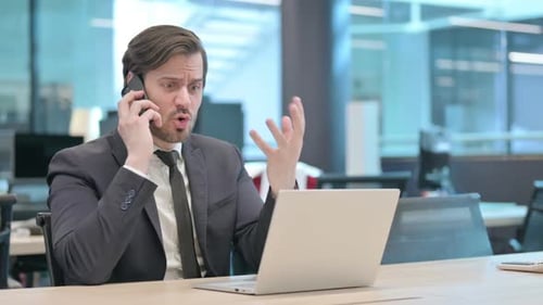 Angry Businessman Talking on Phone While Using Laptop in Office