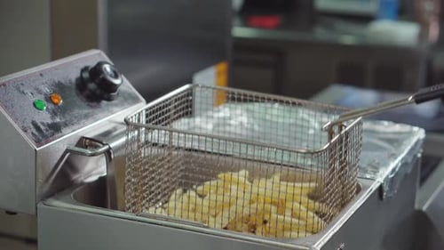 Kitchen Worker Is Pouring Frozen Pieces of Potato Inside Grid of Fryer, Cooking
