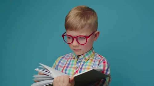 Upset Schoolboy in Brillen liest Buch stehend auf blauem Hintergrund