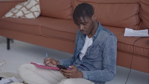 Junger fokussierter Mann, der Idee im Skizzenblock mit Stift schreibt und E-Mail auf dem Smartphone-Bildschirm betrachtet