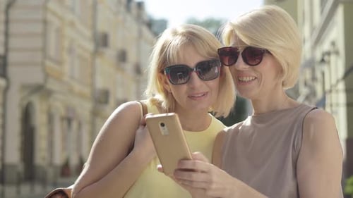 Two Blonde Women Looking at Smartphone in City
