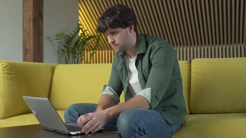 Attractive Blackhaired Guy Is Looking Through His Laptop While Sitting at Home on a Cozy Yellow Sofa