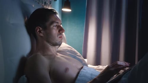 Portrait of Man Freelancer Lying on a Bed in the Bedroom at Night at Home and Working with Laptop