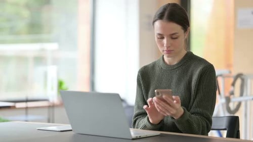 Young Woman Scrolling Through Her Phone at Desk