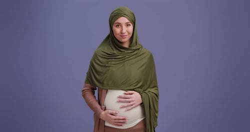 Studio Shot of Pregnant Islamic Lady Stroking Her Big Belly and Smiling to Camera Purple Background