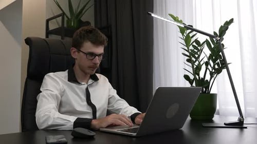 Jovem bonito de óculos e camisa está sentado em casa, escritório, local de trabalho, abrindo laptop