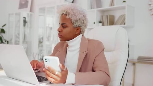 Young African American Business Woman Works with Phone and Laptop Sits in Office