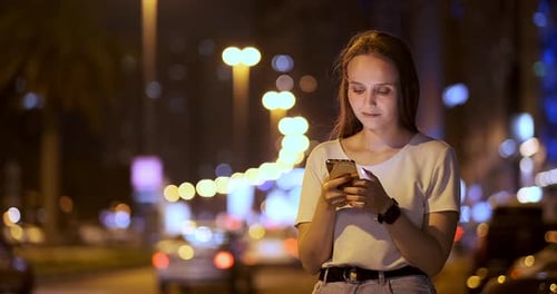 A Young Girl on the Background of the Night City Looks Into the Smartphone Screen