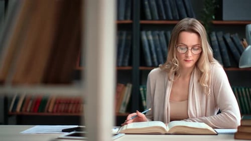 Confident Woman Student Reading Paper Book Prepare to Exam at Public Library Desk Bookshelves