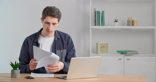Caucasian Male Freelancer Sits at Table in His Home Office Reads Documents Compares Numbers in