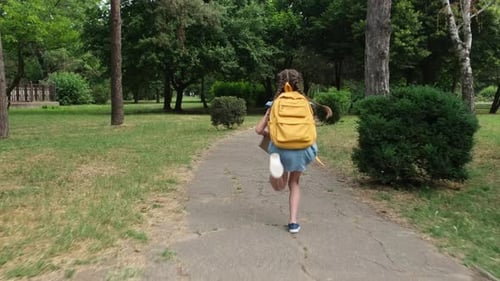 Kleines Schulmädchen mit Schulrucksack und Buch rennt durch den Park zur Schule