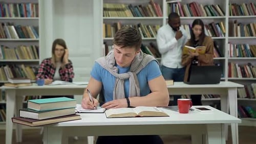 Smart Guy Sitting at the Table in the Library and Doing His University Home Task