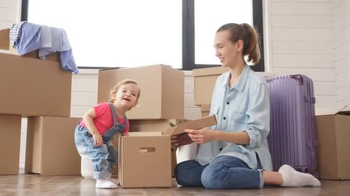 Mom with Her Little Daughter Sitting on Floor in the New House and Dismantle Boxes