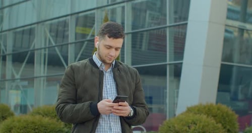 Portrait of Man Walking Near Modern Office Center with Smartphone and Earphones, Chatting in Social
