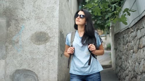 Young Woman with Backpack on Her Shoulder Walking Along in Old European Town in Summer Day