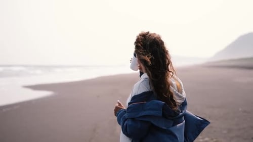 Beautiful Young Woman in the Morning Stands on the Beach with Black Volcanic Sand and Drinks Coffee