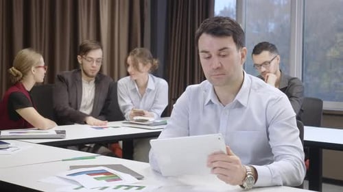 Concentrated Caucasian Man Scrolling Tablet Sitting in Office with Colleagues at Background Looking
