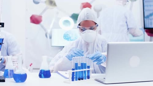 Female Scientist Picking Up a Test Tube with Blue Solution and Typing on Computer
